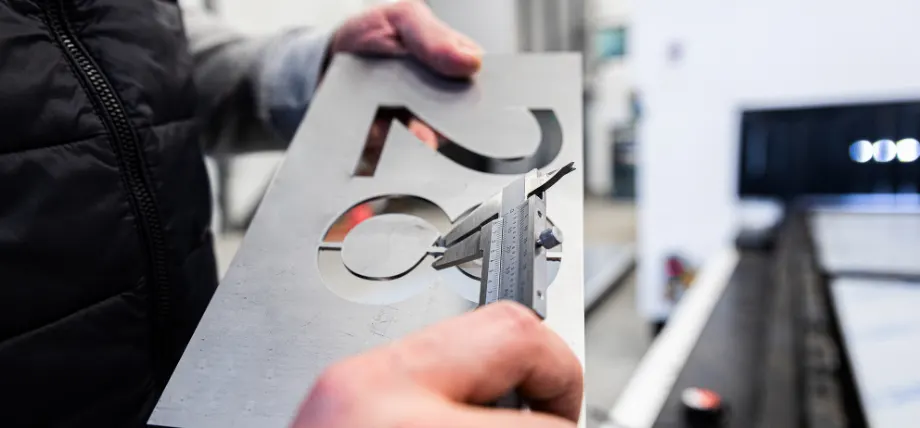 Technician measuring laser-cut metal numbers with digital caliper to verify tight tolerances.